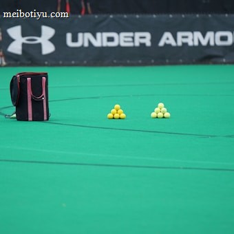 A sports field features a black and pink sports bag, two pyramids of yellow balls, and a portion of a person in dark athletic gear. The background displays a banner with the Under Armour logo.