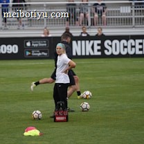 A person in sports attire stands on a soccer field, surrounded by several soccer balls and a carrier with bottles. The field is bordered by advertisements for Nike Soccer and app stores. Several people sit in bleachers in the background.