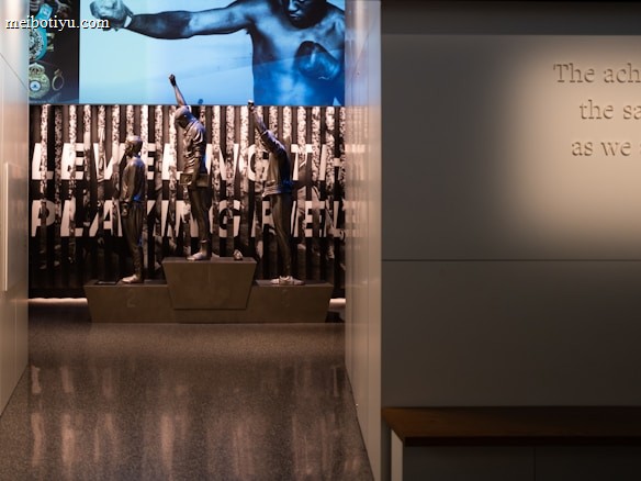 A dimly lit display features statues of athletes standing on a podium. They are positioned in front of a backdrop with the words 'LEVELING THE PLAYING FIELD' in bold letters. On the wall to the right, a softly lit quote is partially visible. The overall ambiance suggests a museum or exhibition setting.