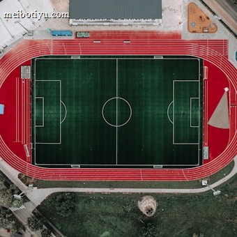 An aerial view of a large sports complex featuring a central green football field surrounded by a red running track. Adjacent to the track, there is a parking area, buildings, and pathways. Nearby, there are additional sports courts in different colors, including a blue swimming pool and orange basketball courts.