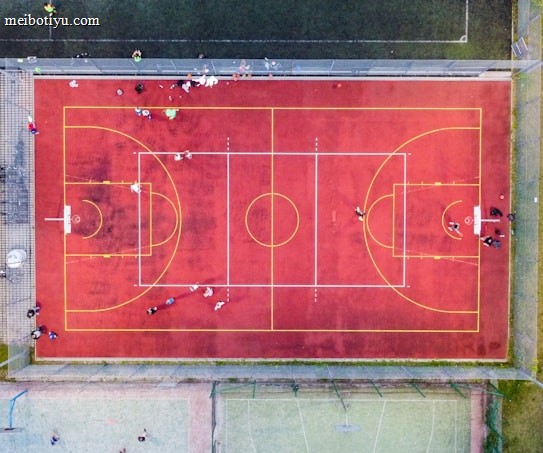An aerial view of a sports court with a vibrant red surface and yellow lines marking different sections for basketball and soccer. Several people are present on the court, some engaged in activities, while others are standing along the perimeter. The court is enclosed by a metal fence and surrounded by grass on all sides.